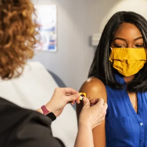 Photo: a health care provider and patient wearing a face mask, inside a clinical setting, the provider is placing a colorful bandage of the vaccination shot site on the patient's upper arm. (Source: CDC, unsplash)